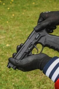 Close-up image showing hands with gloves holding a pistol in a grassy area.